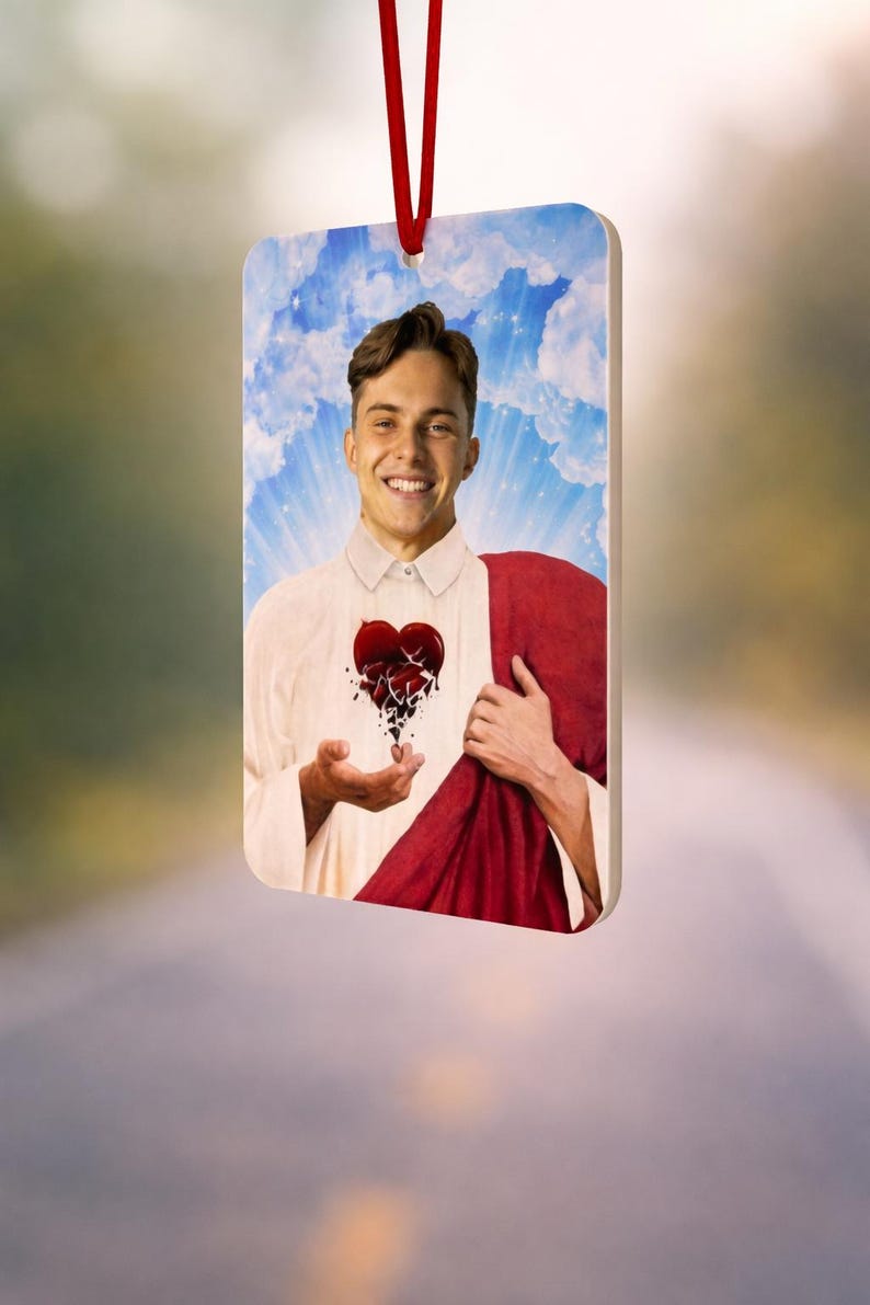 May include: Rectangular ornament with a smiling person and a broken heart design. The person wears a white shirt and red cloth. The background shows a blue sky with clouds. A red ribbon is attached for hanging.