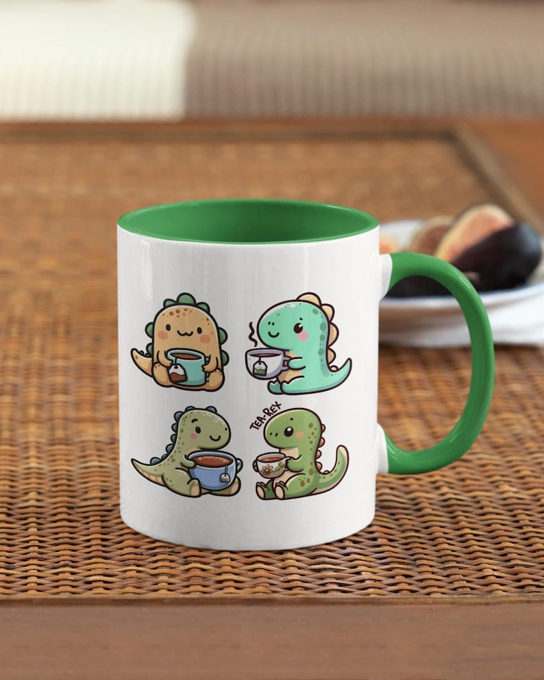 May include: White ceramic mug with a green rim and handle. The mug features four illustrations of cartoon dinosaurs drinking tea or coffee. The text "Tea-Rex" is printed on the mug.