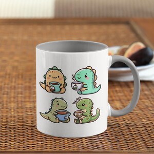 May include: A white ceramic mug with a gray rim and handle. The mug features a cartoon illustration of four dinosaurs drinking tea. The text "Tea Rex" is printed on the mug.