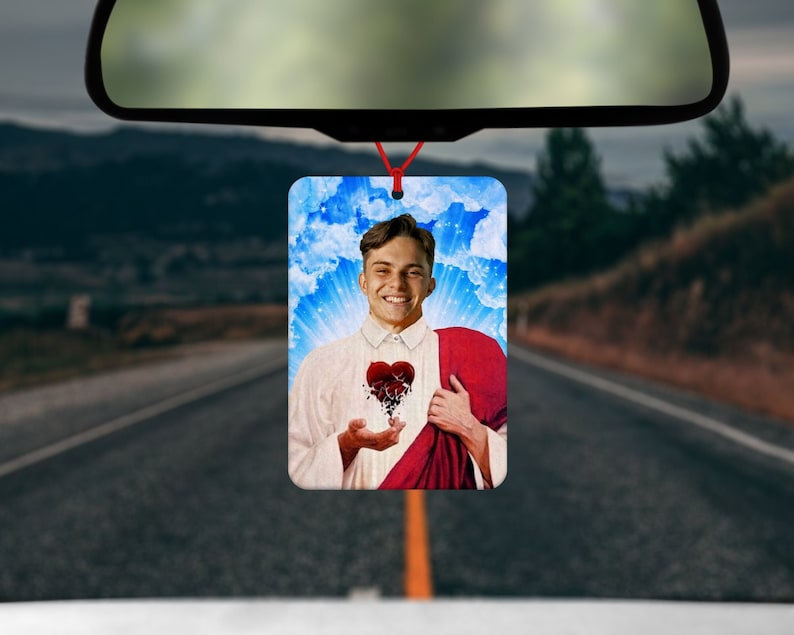 May include: Rectangular car air freshener featuring a smiling person with a broken heart design. The background includes a blue sky with clouds and sun rays. The air freshener hangs from a red string.