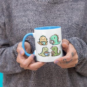 May include: White ceramic mug with a blue rim. The mug features a cartoon illustration of four dinosaurs drinking tea. The dinosaurs are green and yellow.