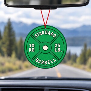 May include: A circular green air freshener hanging from a rearview mirror by a red string. The design mimics a weight plate with the text STANDARD BARBELL, 10 KG, and 25 LB. printed in white, featuring a central hole and four spokes.