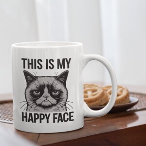 May include: White ceramic mug with a black and white illustration of a grumpy cat with the text "This is my happy face" printed on it.