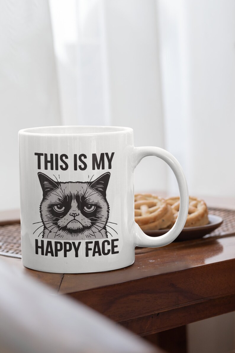 May include: White ceramic mug with a black and white illustration of a grumpy cat with the text "This is my happy face" printed on it.