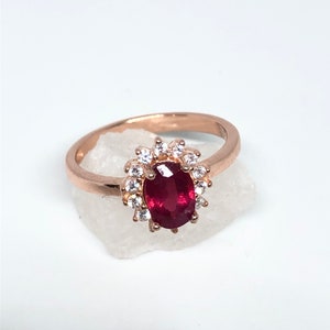 May include: A rose gold ring with a large oval ruby gemstone surrounded by a halo of smaller white gemstones.
