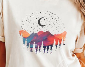 May include: A cream-coloured t-shirt with a watercolour design of a mountain range, trees, a crescent moon, and stars. The mountains are in shades of red, purple, and blue. The trees are dark green, and the moon and stars are black.