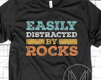 Puede incluir: Camiseta negra con el texto "EASILY DISTRACTED BY ROCKS" en verde azulado, blanco, amarillo y naranja. La camiseta tiene cuello redondo y mangas cortas. Un logotipo es visible en la parte inferior derecha.