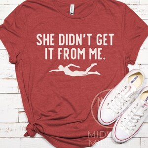 May include: Red t-shirt with the text "She didn't get it from me." and a white silhouette of a swimmer.