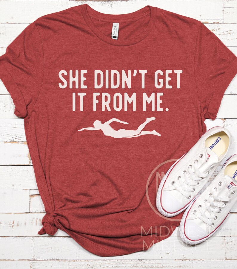 May include: Red t-shirt with the text "She didn't get it from me." and a white silhouette of a swimmer.