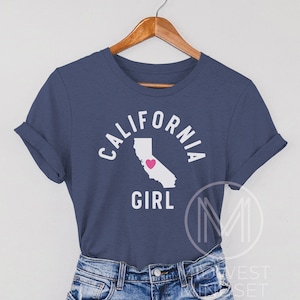Pu&ograve; includere: Una maglietta blu navy con il testo "CALIFORNIA GIRL" in bianco con un contorno bianco dello stato della California con un cuore rosa al centro.