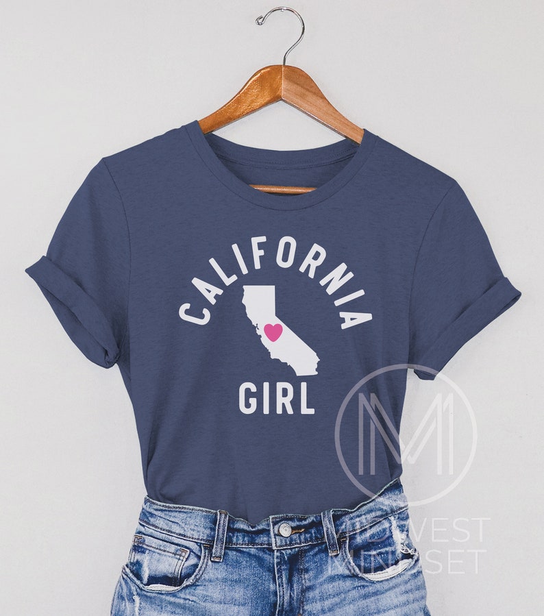 Pu&ograve; includere: Una maglietta blu navy con il testo "CALIFORNIA GIRL" in bianco con un contorno bianco dello stato della California con un cuore rosa al centro.