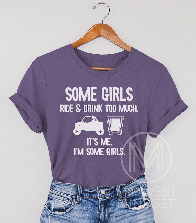 May include: Purple t-shirt with white text that reads "Some girls ride & drink too much. It's me. I'm some girls."  The shirt features a white silhouette of an ATV and a shot glass.
