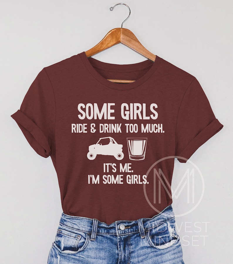 May include: A maroon t-shirt with white text that reads "Some girls ride & drink too much. It's me. I'm some girls."  The text is accompanied by a graphic of a side-by-side vehicle and a shot glass.