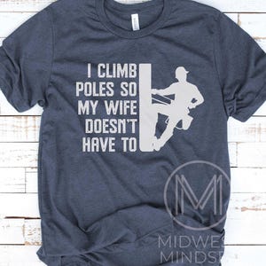 Puede incluir: Una camiseta azul marino con un gr&aacute;fico blanco de un lineman escalando un poste de electricidad. El texto en la camiseta dice "Subo a los postes para que mi esposa no tenga que hacerlo."