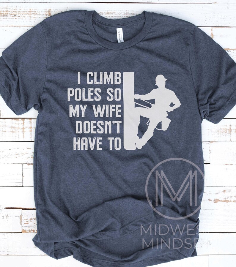 Puede incluir: Una camiseta azul marino con un gr&aacute;fico blanco de un lineman escalando un poste de electricidad. El texto en la camiseta dice "Subo a los postes para que mi esposa no tenga que hacerlo."
