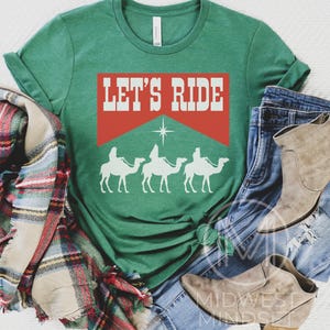 Pu&ograve; includere: Una maglietta verde con il testo "Let's Ride" in lettere bianche su uno sfondo rosso. Sotto il testo ci sono tre sagome bianche di persone che cavalcano cammelli.