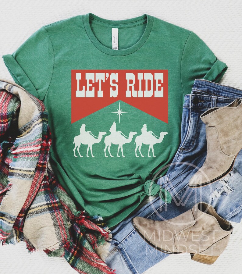 Pu&ograve; includere: Una maglietta verde con il testo "Let's Ride" in lettere bianche su uno sfondo rosso. Sotto il testo ci sono tre sagome bianche di persone che cavalcano cammelli.