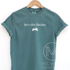 Puede incluir: Camiseta de color verde azulado con la frase "let's ride bitches" en blanco y un peque&ntilde;o gr&aacute;fico de un coche. La camiseta se muestra en una percha de madera sobre un fondo blanco. Las mangas est&aacute;n dobladas.