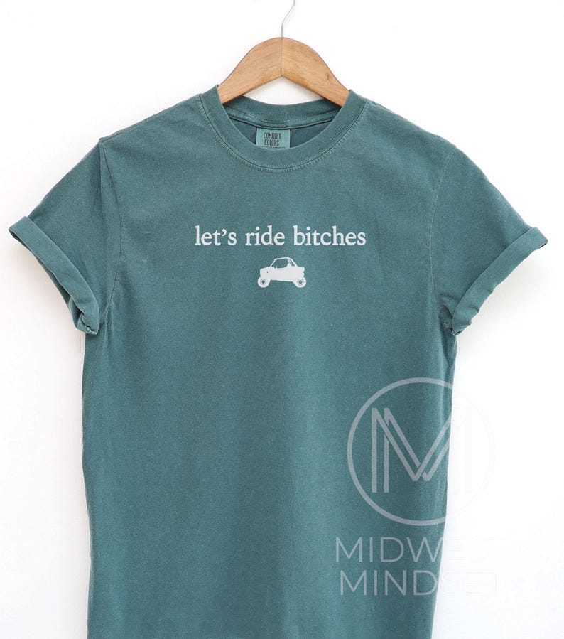Puede incluir: Camiseta de color verde azulado con la frase "let's ride bitches" en blanco y un peque&ntilde;o gr&aacute;fico de un coche. La camiseta se muestra en una percha de madera sobre un fondo blanco. Las mangas est&aacute;n dobladas.