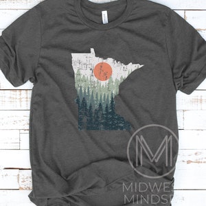 May include: A dark grey t-shirt with a distressed graphic of the state of Minnesota. The graphic features a forest scene with a large orange sun setting behind the trees.