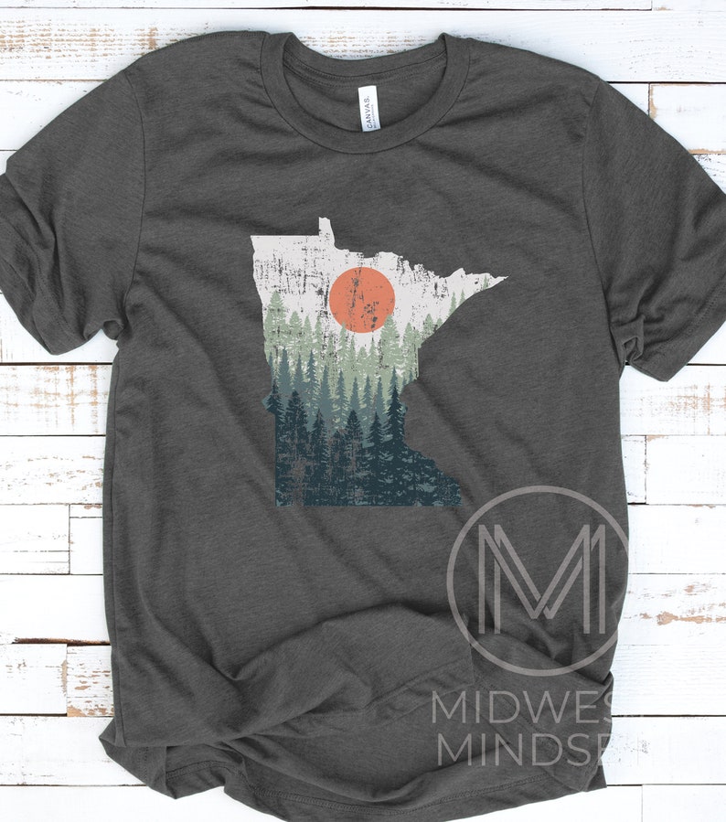 May include: A dark grey t-shirt with a distressed graphic of the state of Minnesota. The graphic features a forest scene with a large orange sun setting behind the trees.