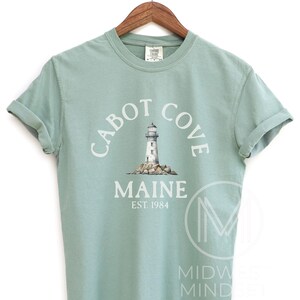 以下が含まれることがあります： 白文字で「CABOT COVE MAINE EST. 1984」と灯台のイラストが描かれた、淡い緑色のTシャツ。木製のハンガーに掛けられています。