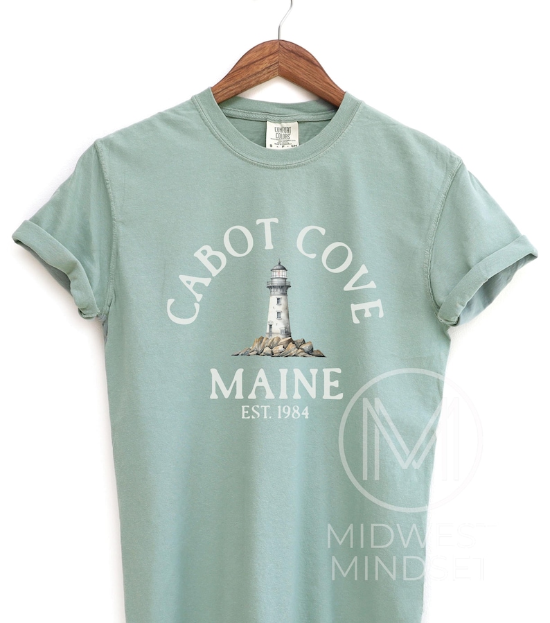 以下が含まれることがあります： 白文字で「CABOT COVE MAINE EST. 1984」と灯台のイラストが描かれた、淡い緑色のTシャツ。木製のハンガーに掛けられています。