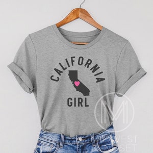 Pu&ograve; includere: Maglietta grigia con un contorno nero dello stato della California e un cuore rosa al centro. Il testo "California Girl" &egrave; stampato sotto il contorno dello stato.