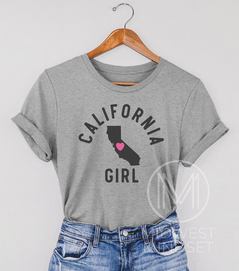Pu&ograve; includere: Maglietta grigia con un contorno nero dello stato della California e un cuore rosa al centro. Il testo "California Girl" &egrave; stampato sotto il contorno dello stato.