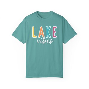 Puede incluir: Camiseta turquesa con la frase "LAKE vibes". La palabra "LAKE" est&aacute; en letras de bloque amarillas, naranjas, rosas y azules, con "vibes" en cursiva. Una camiseta informal y c&oacute;moda.