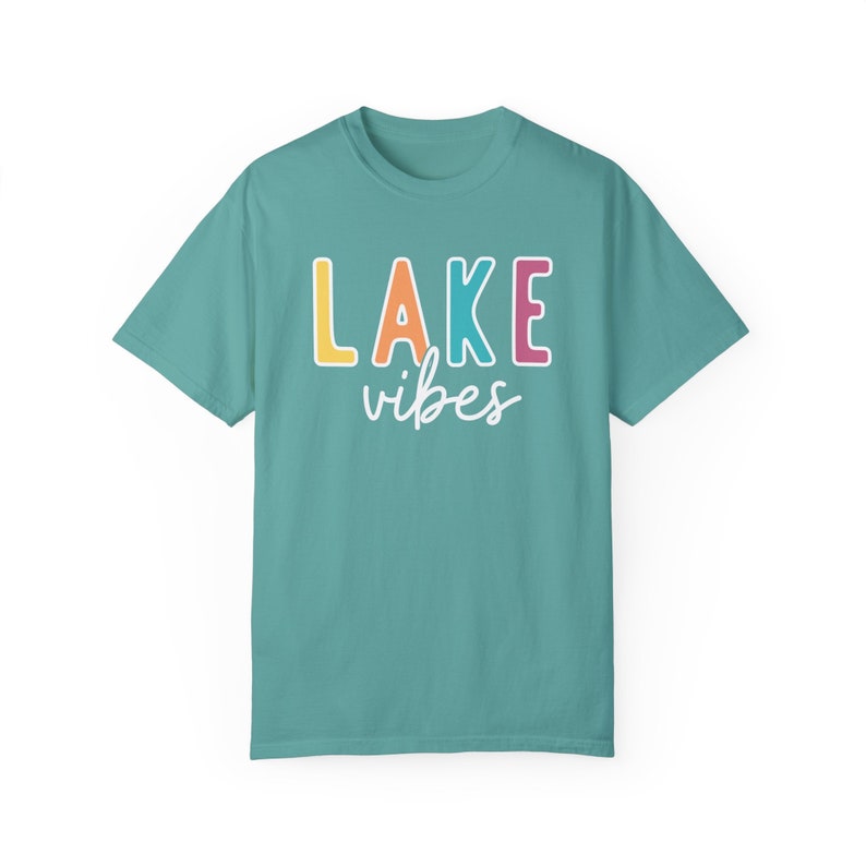 Puede incluir: Camiseta turquesa con la frase "LAKE vibes". La palabra "LAKE" est&aacute; en letras de bloque amarillas, naranjas, rosas y azules, con "vibes" en cursiva. Una camiseta informal y c&oacute;moda.