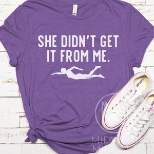 May include: Purple t-shirt with the text "She didn't get it from me." and a white silhouette of a swimmer.