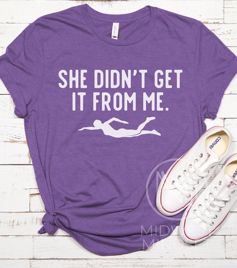 May include: Purple t-shirt with the text "She didn't get it from me." and a white silhouette of a swimmer.