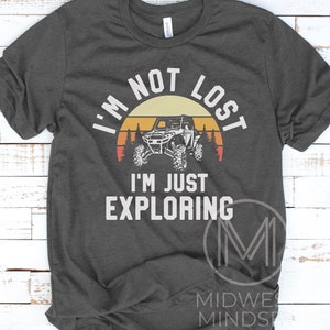K&ouml;nnte beinhalten: Ein dunkelgraues T-Shirt mit einer Grafik im Vintage-Stil eines Sonnenuntergangs, B&auml;umen und einem Offroad-Fahrzeug mit zwei Sitzen. Der Text "Ich bin nicht verloren, ich erkunde nur" ist auf das T-Shirt gedruckt.