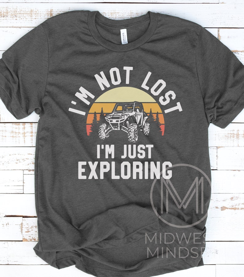 K&ouml;nnte beinhalten: Ein dunkelgraues T-Shirt mit einer Grafik im Vintage-Stil eines Sonnenuntergangs, B&auml;umen und einem Offroad-Fahrzeug mit zwei Sitzen. Der Text "Ich bin nicht verloren, ich erkunde nur" ist auf das T-Shirt gedruckt.