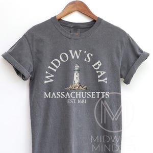 May include: A charcoal gray short-sleeve t-shirt featuring a graphic of a lighthouse on rocks. The text reads WIDOW'S BAY, MASSACHUSETTS, and EST. 1681. The shirt is displayed on a wooden hanger, showcasing a crew neckline and rolled sleeve cuffs.