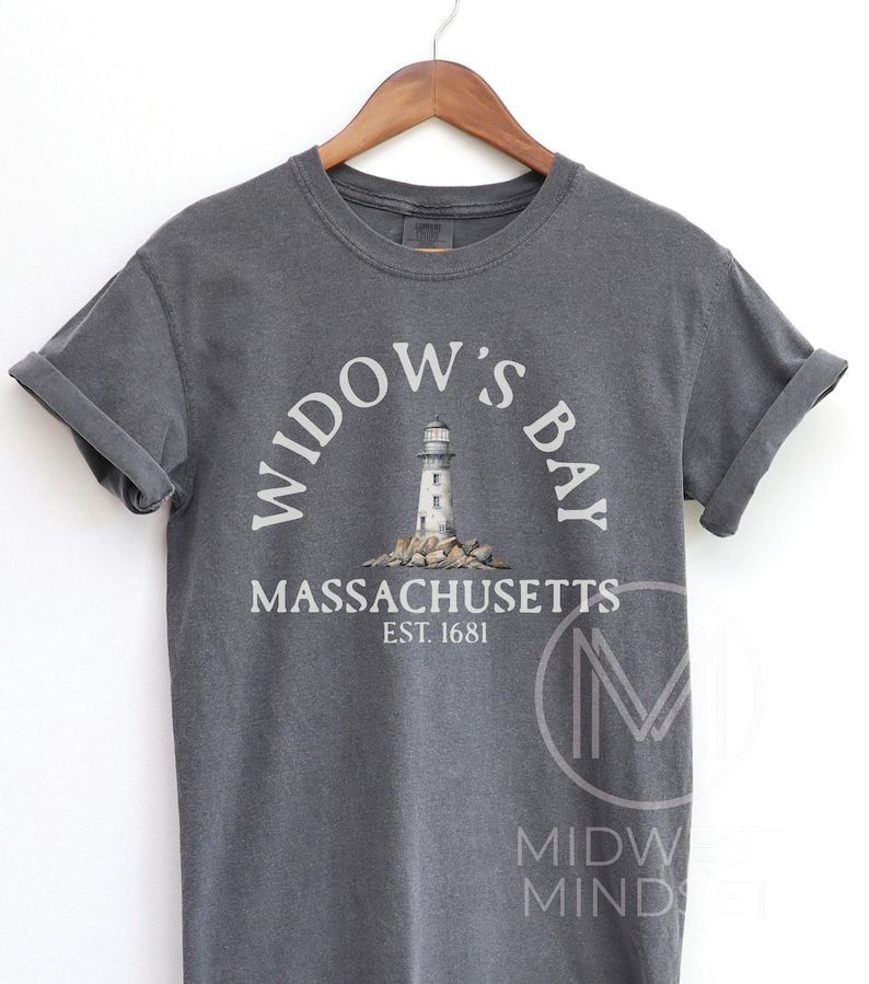 May include: A charcoal gray short-sleeve t-shirt featuring a graphic of a lighthouse on rocks. The text reads WIDOW'S BAY, MASSACHUSETTS, and EST. 1681. The shirt is displayed on a wooden hanger, showcasing a crew neckline and rolled sleeve cuffs.