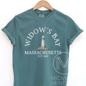 May include: A teal short-sleeve t-shirt featuring a graphic of a lighthouse on rocks. The text reads WIDOW'S BAY, MASSACHUSETTS, EST. 1681 in white lettering. The shirt is displayed on a wooden hanger, showcasing a crew neckline and rolled sleeves.
