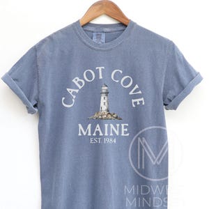 以下が含まれることがあります： 「Cabot Cove Maine」の文字と灯台のグラフィックがプリントされた青いTシャツ。 「EST. 1984」の文字が「Maine」の下にあります。木製のハンガーにTシャツがかけられています。
