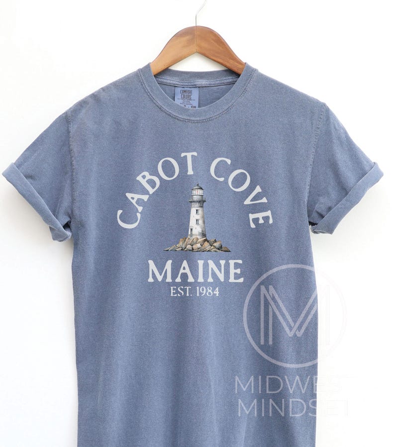 以下が含まれることがあります： 「Cabot Cove Maine」の文字と灯台のグラフィックがプリントされた青いTシャツ。 「EST. 1984」の文字が「Maine」の下にあります。木製のハンガーにTシャツがかけられています。
