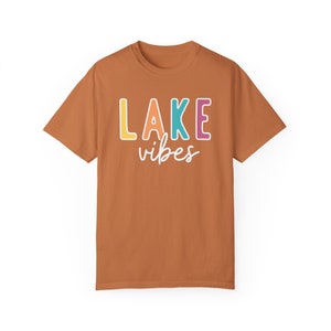 Puede incluir: Camiseta de color &oacute;xido con la frase "LAKE vibes" estampada. La palabra "LAKE" est&aacute; en letras de bloque coloridas, y "vibes" en cursiva. La camiseta es de manga corta y cuello redondo, perfecta para el verano.
