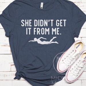 May include: A navy blue t-shirt with the text "She didn't get it from me." in white. A white silhouette of a swimmer is below the text.