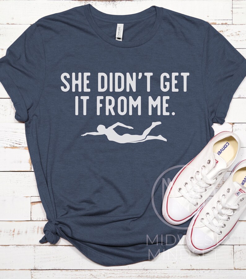 May include: A navy blue t-shirt with the text "She didn't get it from me." in white. A white silhouette of a swimmer is below the text.