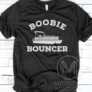May include: A dark gray t-shirt with the text "Boobie Bouncer" in white. A white silhouette of a pontoon boat is below the text. The text "MIDWEST MINDSET" is in white on a black M logo in the lower right corner.