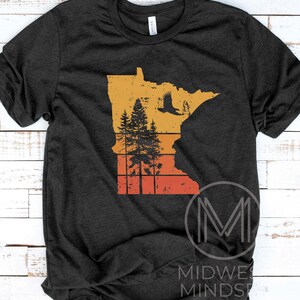 May include: A dark grey t-shirt with a vintage-style graphic of the state of Minnesota. The graphic features a silhouette of a soaring eagle, pine trees, and a sunset-colored background.