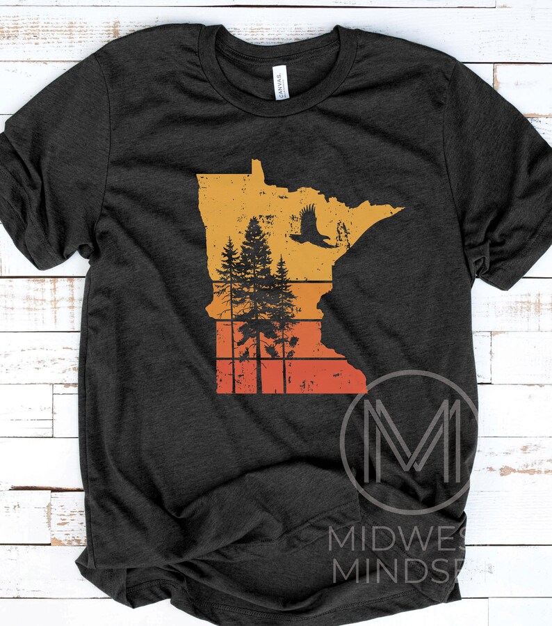 May include: A dark grey t-shirt with a vintage-style graphic of the state of Minnesota. The graphic features a silhouette of a soaring eagle, pine trees, and a sunset-colored background.