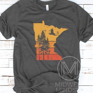 May include: A dark gray t-shirt with a vintage-style graphic of the state of Minnesota. The graphic features a silhouette of a forest with an eagle flying overhead, all set against a sunset background.