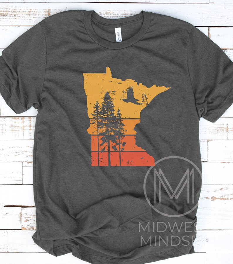 May include: A dark gray t-shirt with a vintage-style graphic of the state of Minnesota. The graphic features a silhouette of a forest with an eagle flying overhead, all set against a sunset background.