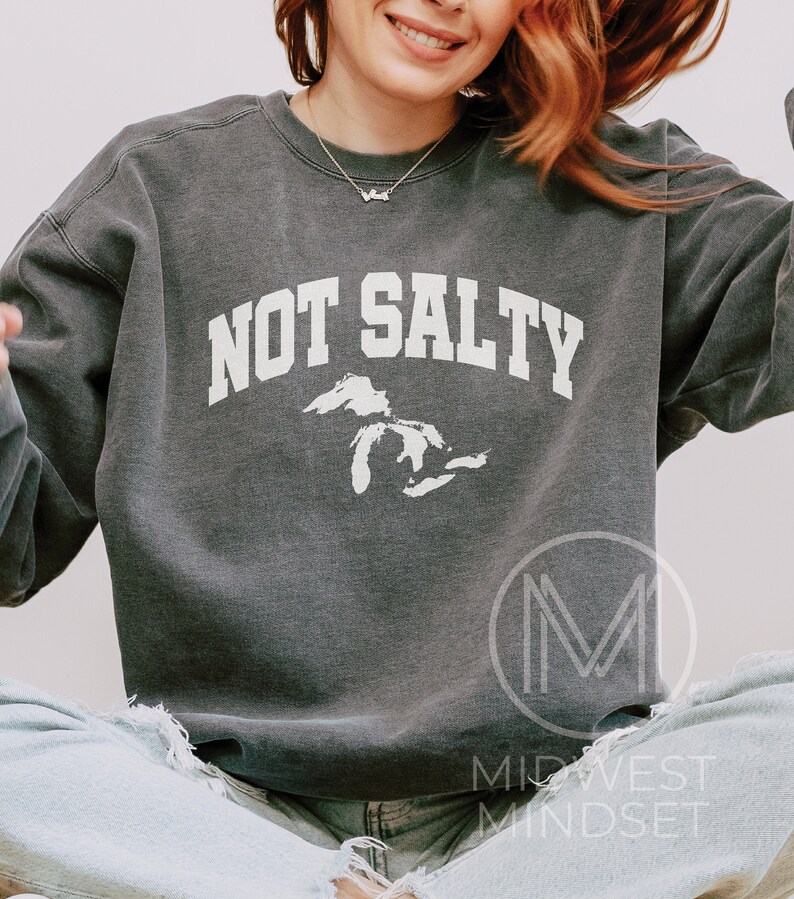 May include: A dark gray sweatshirt with the text "NOT SALTY" in white, with a white outline of the Great Lakes. The sweatshirt is worn by a person sitting with their legs crossed.