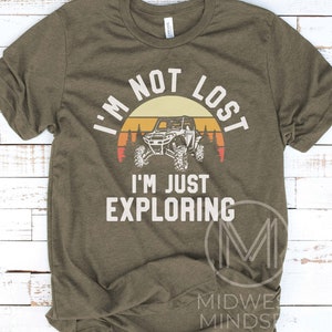 K&ouml;nnte beinhalten: Ein gr&uuml;nes T-Shirt mit einer Retro-Sonnenuntergangsgrafik und einer Silhouette eines Gel&auml;ndewagens. Der Text "Ich bin nicht verloren, ich erkunde nur" ist auf dem Shirt aufgedruckt.